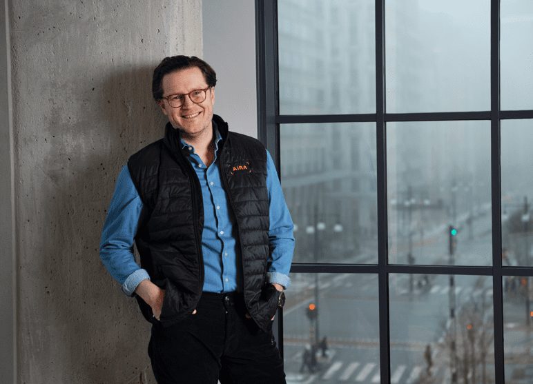 A man from Aira in a padded gilet, leaning against a window with a city view.