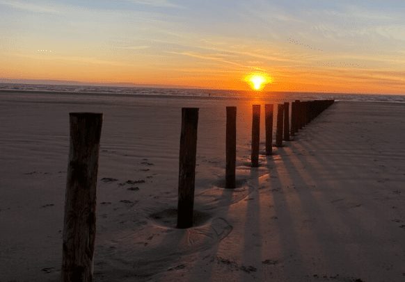 Sunset over the sands beside a holiday park, with a line of wooden posts on the beach.