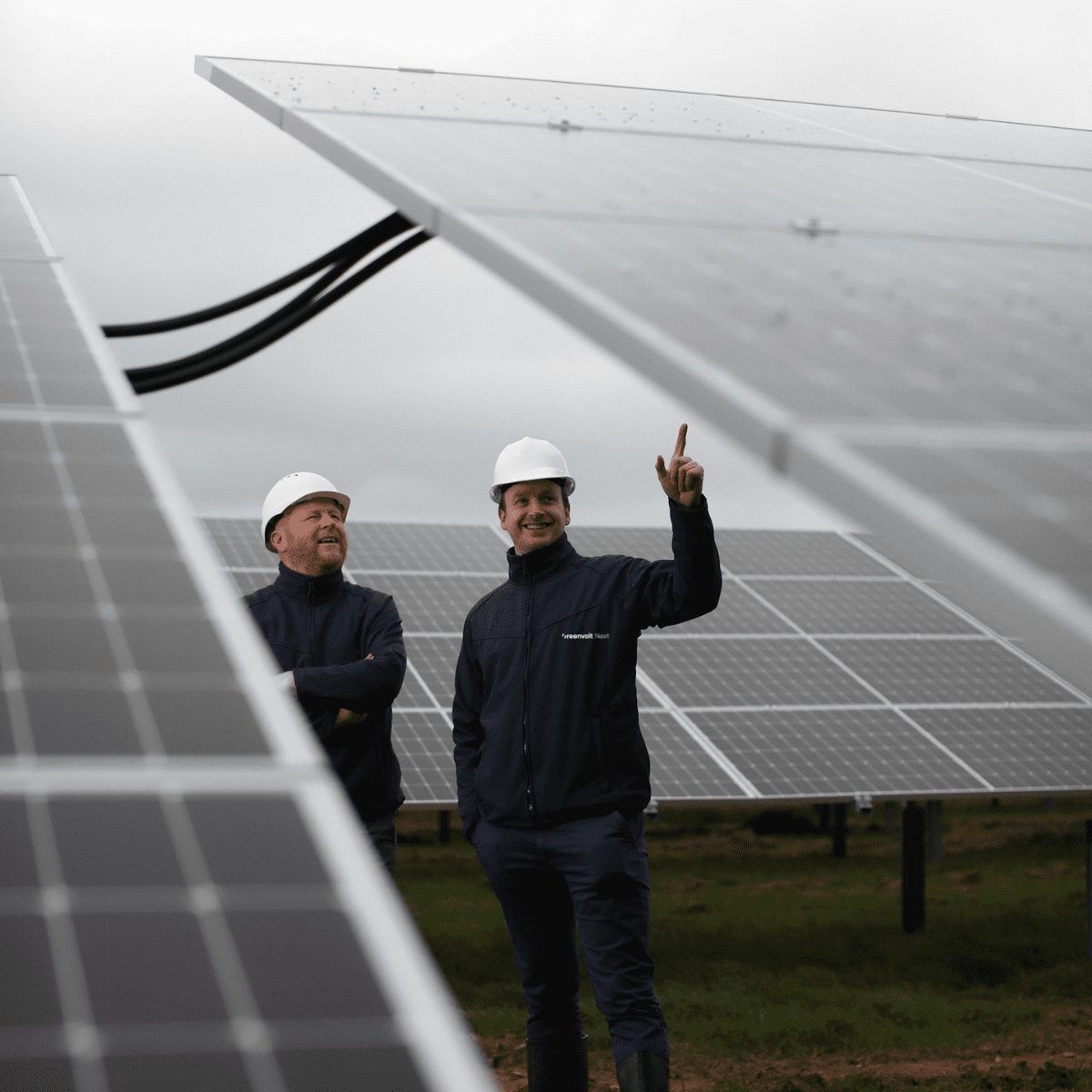Two men in hard hats beneath solar panels at a solar farm, one pointing upwards.