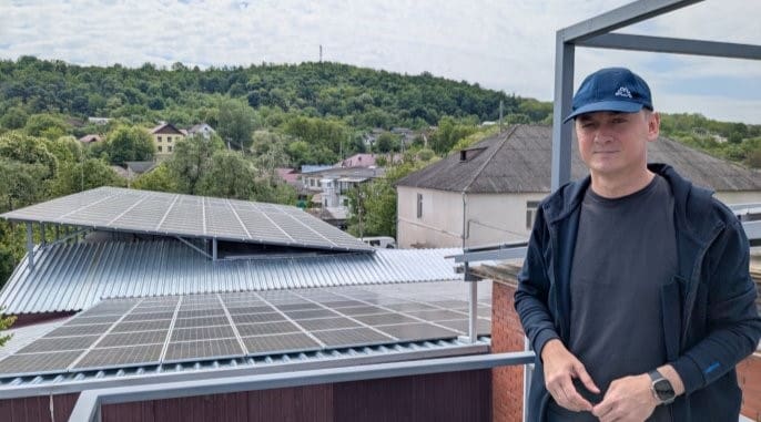 A man in a cap stands on a rooftop beside an angled array of solar panels, with houses and wooded hills behind.