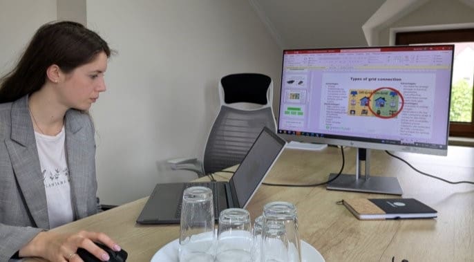 A woman at a desk looks at a presentation slide on a large monitor, with a laptop and water glasses in front of her.