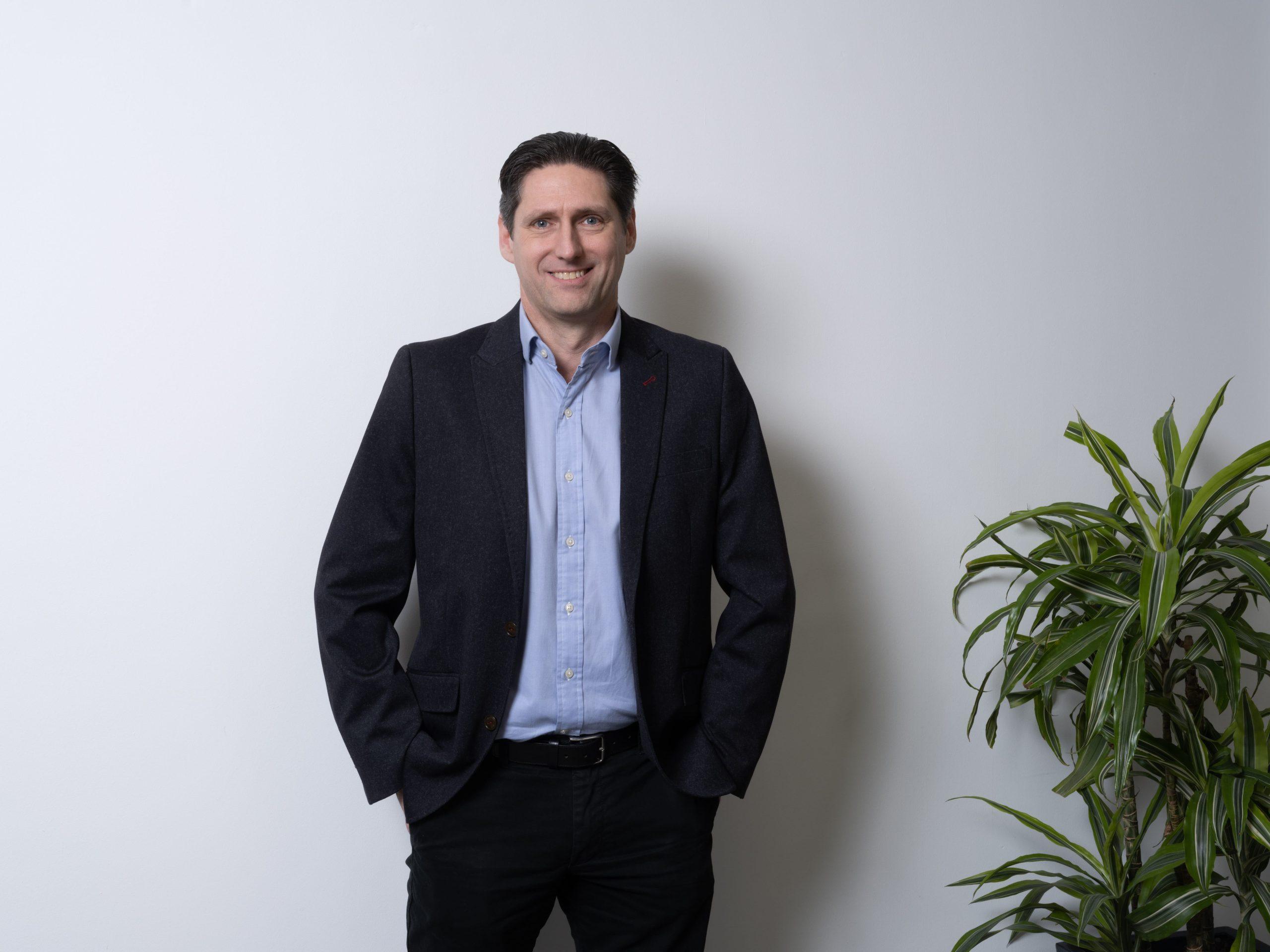 Paul Rogers, in a dark suit and open-necked shirt, standing beside a plant against a white wall.