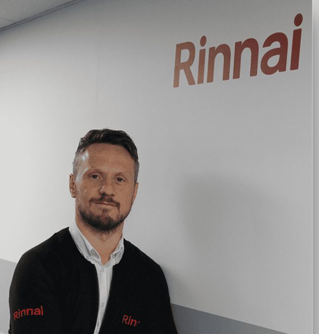 Chris Goggin of Rinnai, in a dark Rinnai-branded top, in front of a wall with the Rinnai logo.