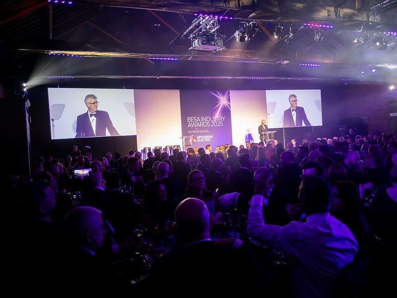A presenter on stage at the BESA Awards, in front of a large audience and screens.