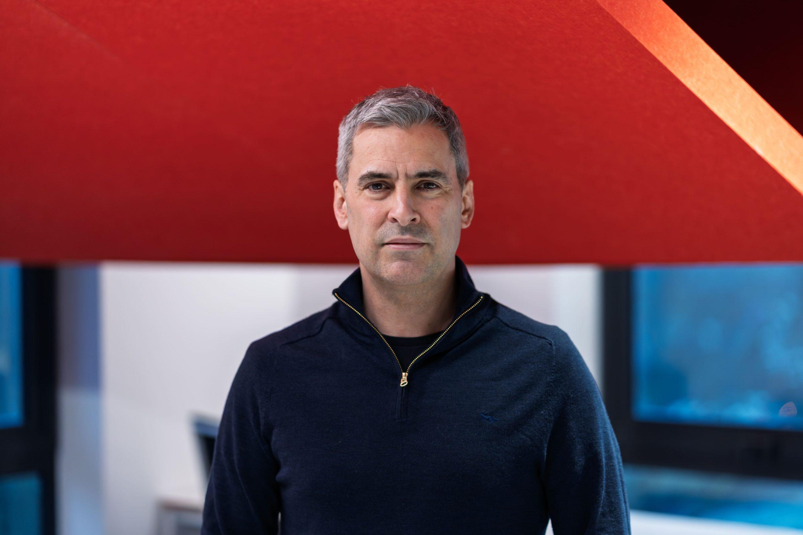 Christophe Williams in a dark jumper in front of a red and white background.