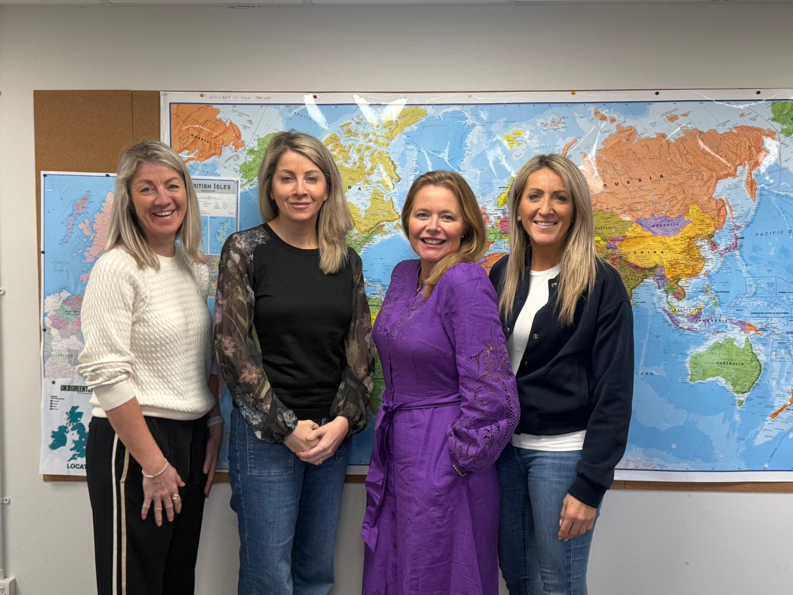 Four women stand in front of a world map in an office.