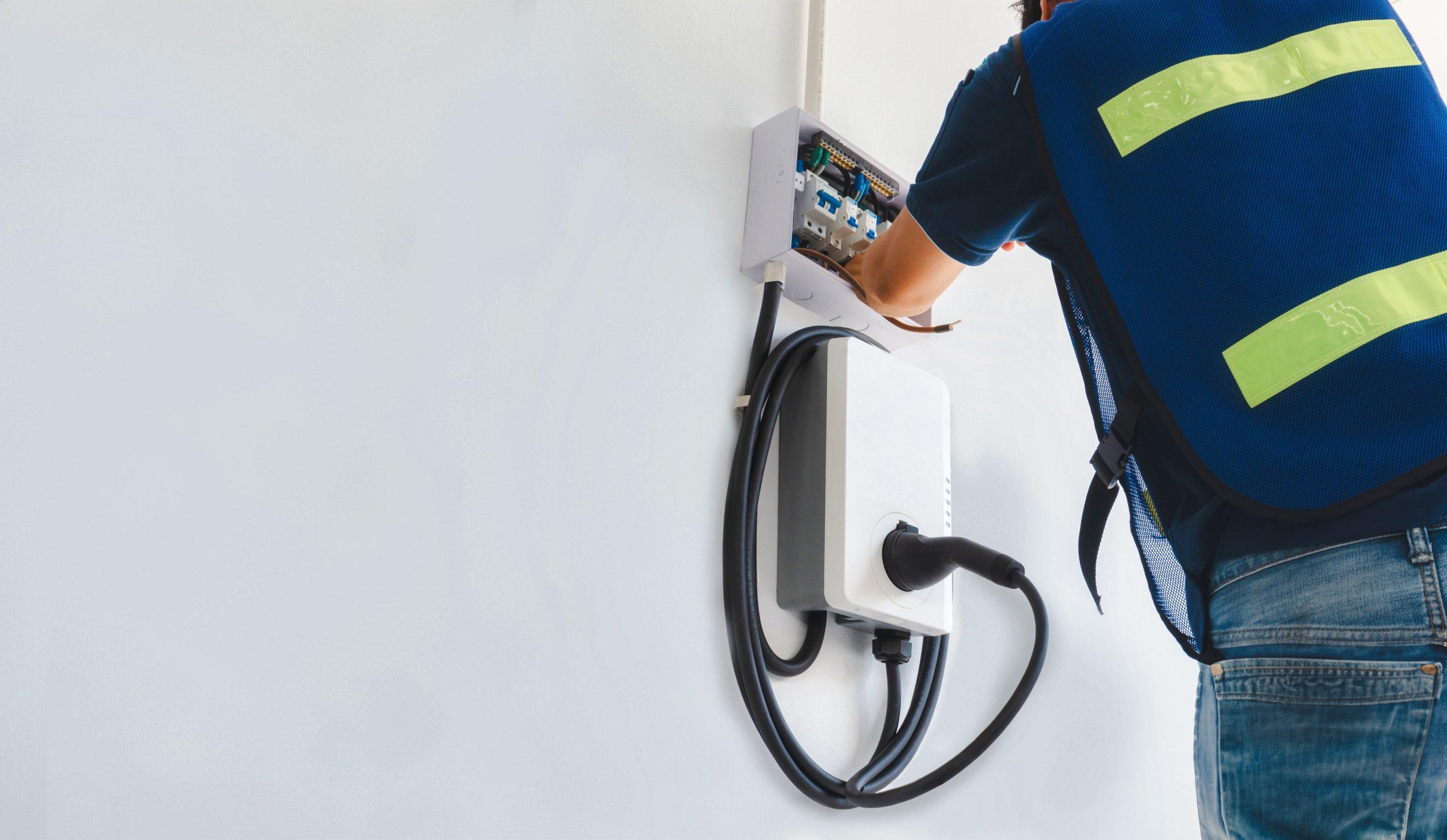 An electrician in a hi-vis vest installs a home EV charger on a white wall.