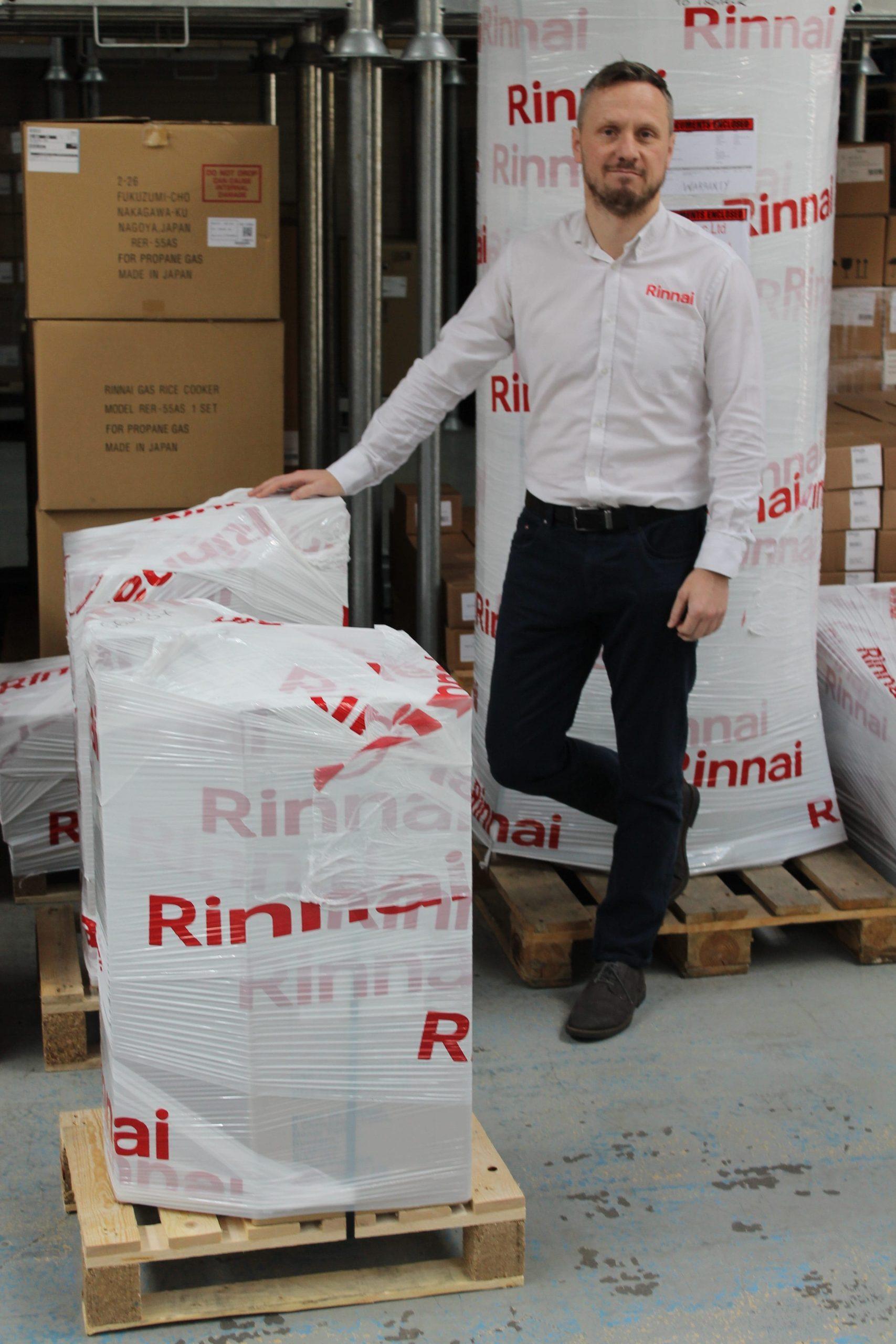 A man in a white shirt stands among stacked, wrapped Rinnai units on pallets in a warehouse.