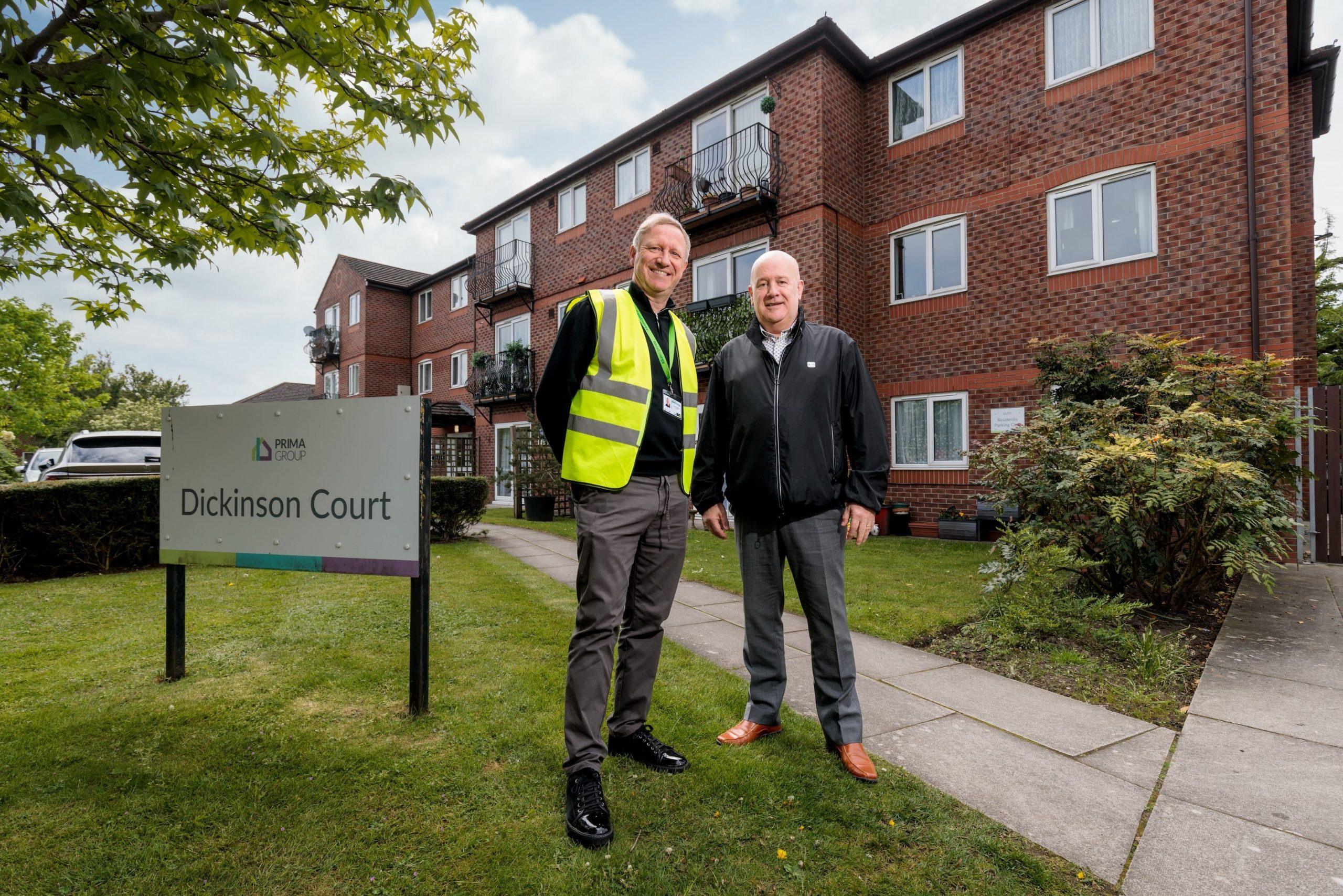 Two men, one in a hi-vis vest, stand by the Dickinson Court sign in front of a red-brick block of flats.