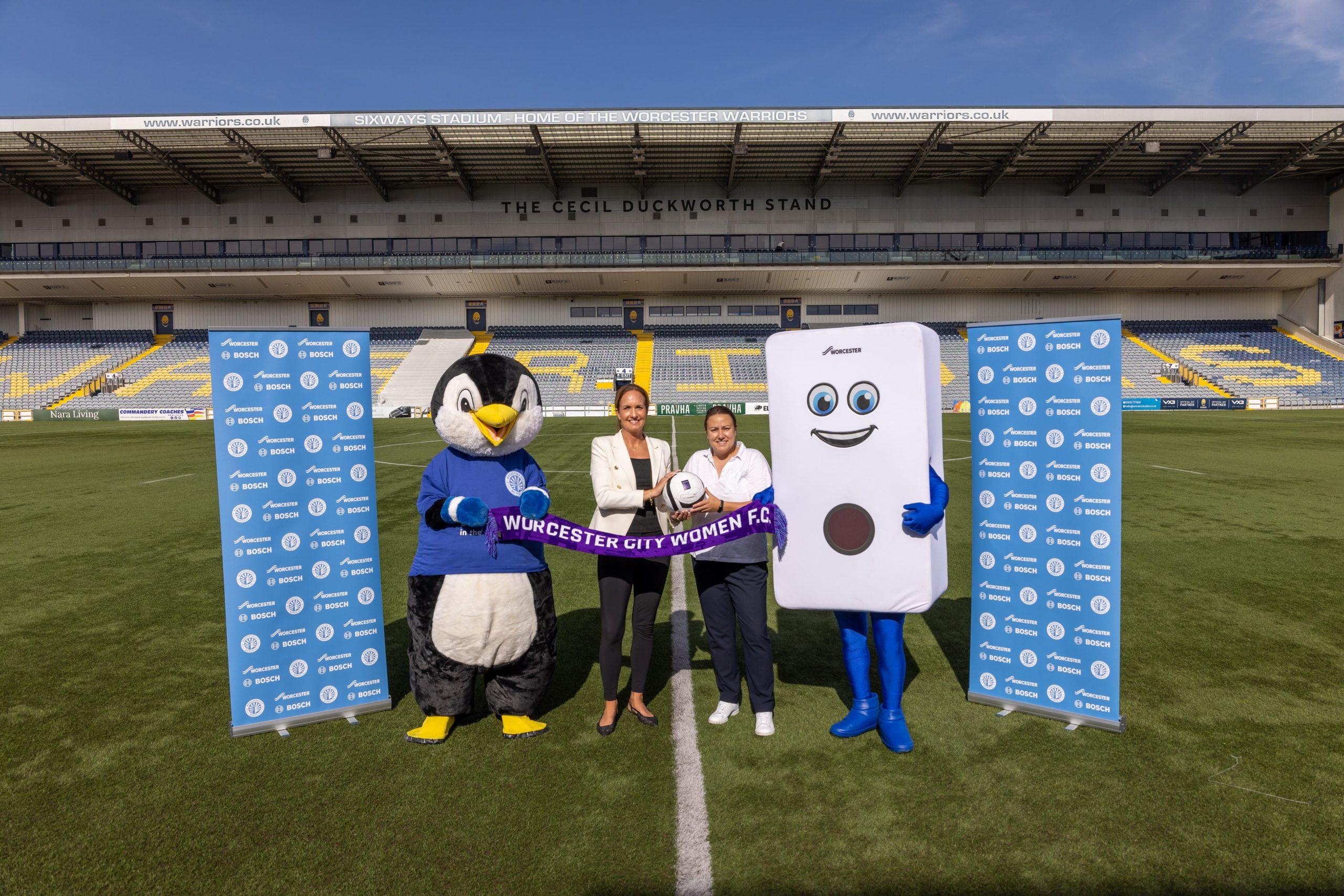 Two club mascots and two people stand on a rugby pitch between Worcester Bosch banners at a stadium.