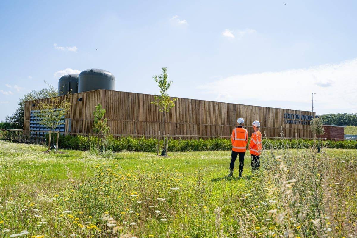 Two people in hi-vis jackets stand in a wildflower meadow in front of a timber-clad Community Heat Hub building at Chilton Woods.